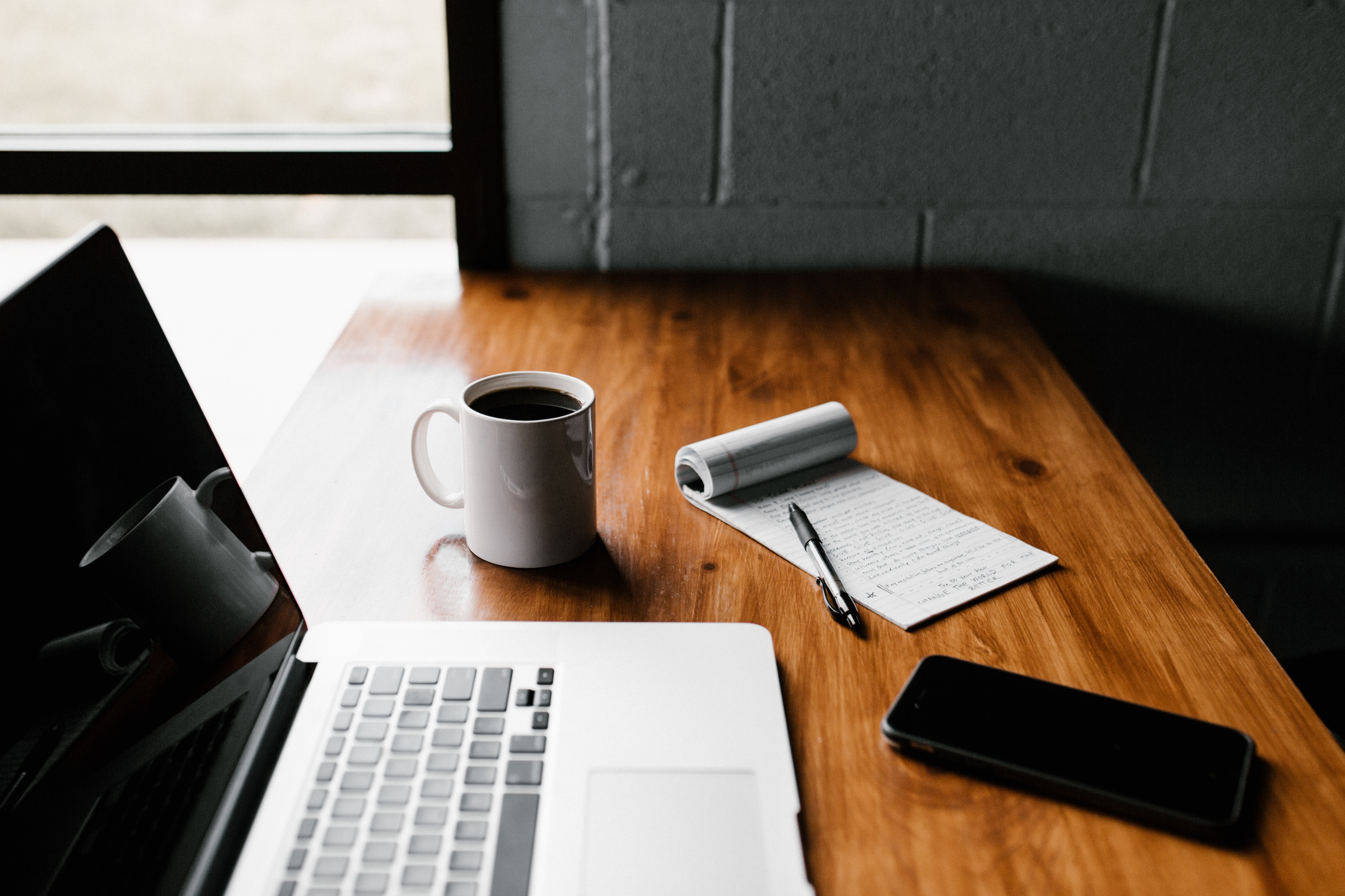 Macbook, phone, coffee, and paper pad on a wooden desk